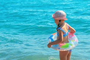 un' poco ragazza con un gonfiabile squillare nel un' costume da bagno e un' sole cappello entra il onde di il oceano su un' soleggiato caldo giorno foto