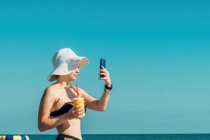 giovane bellissimo donna nel un' sole cappello e costume da bagno, Tenere un' bicchiere con un' freddo estate bevanda nel sua mani, prende un' autoscatto su il Telefono mentre rilassante su il oceano spiaggia foto