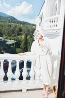 ritratto donna nel un' bianca accappatoio su il balcone po su verde natura Perfetto soleggiato mattina foto