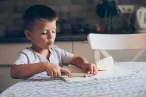 carino ragazzo 4-5 anni vecchio con coltello taglio un' formaggio su il taglio tavola su il tavolo nel il cucina foto