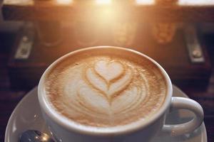 vicino su di un' tazza di caldo caffè latte macchiato su di legno tavolo con un' caffè fagioli nel bicchiere tubo a il sfondo. foto