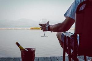 uomo seduta su un' sedia con mano Tenere bicchiere di rosso vino di il lago. foto