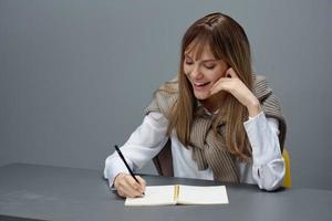 godendo scrittura articolo giovane bionda alunno signora giornalista nel caldo maglione utilizzando quaderno seduta a posto di lavoro nel grigio casa ufficio. a distanza lavoro, formazione scolastica e carriera professione concetto. copia spazio foto