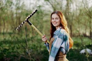 un' donna sorridente meravigliosamente e guardare a il telecamera, un' contadino nel opera Abiti e un grembiule Lavorando all'aperto nel natura e Tenere un' rastrello per raccogliere erba e foraggio per il animali nel il giardino foto