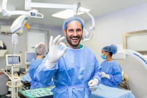 ritratto di maschio chirurgo nel operazione Teatro guardare a telecamera mostrando ok gesto. medico nel scrub e medico maschera nel moderno ospedale operativo camera. foto