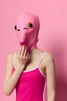 un' divertente donna indossare un' rosa pesce maschera su sua testa mettere sua dita nel sua bocca con rosa Abiti su un' rosa sfondo. il concetto di moderno arte fotografia foto