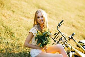 ritratto di un' bellissimo ragazza nel un' campo, seduta su il erba, Il prossimo per un' bicicletta, con un' mazzo di verbena fiori nel il ambientazione sole nel estate foto