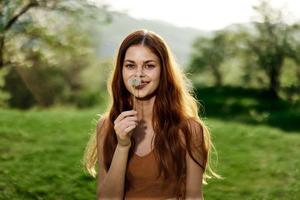 ritratto di un' giovane donna con un' dente di leone fiore nel sua mano soffiaggio su esso e sorridente contro il verde estate erba nel il raggi di il ambientazione sole nel natura foto