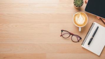 di legno scrivania posto di lavoro con taccuino, penna, occhiali, tavoletta e tazza di caffè, superiore Visualizza piatto posare con copia spazio. foto
