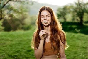 ritratto di un' giovane donna con un' dente di leone fiore nel sua mano soffiaggio su esso e sorridente contro il verde estate erba nel il raggi di il ambientazione sole nel natura foto