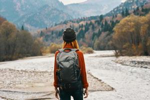 gioioso donna turista con un' zaino sembra a il montagne vicino il fiume su il banca foto