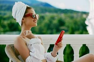 ritratto di giovane donna nel occhiali da sole si siede su il balcone e usi il Telefono Perfetto soleggiato mattina foto