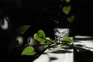 un bicchiere d'acqua su un tavolo bianco con un ramo di una foglia verde. schizza nei raggi del sole. un bicchiere d'acqua foto