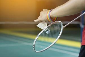 badminton giocatore indossa arcobaleno braccialetti e Tenere racchetta e bianca volano nel davanti di il netto prima servendo esso per giocatore nel un altro lato di il Tribunale, concetto per lgbt persone attività. foto