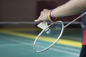 badminton giocatore indossa arcobaleno braccialetti e Tenere racchetta e bianca volano nel davanti di il netto prima servendo esso per giocatore nel un altro lato di il Tribunale, concetto per lgbt persone attività. foto