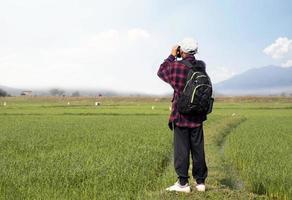 asiatico ragazzo indossare plaid camicia e un' berretto Tenere un' binocolo e guardare a il gregge di uccelli alimentazione su il risaia campo e volante nel il cielo nel il risaia campo. foto