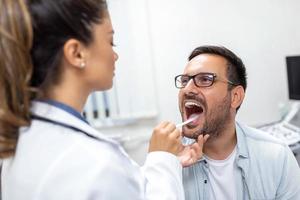 un' giovane uomo si siede su un esame tavolo attraverso a partire dal il suo medico. il medico raggiunge inoltrare con un' lingua depressore come il uomo sembra su e bastoni su il suo lingua. foto