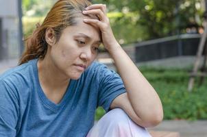 triste e stressato asiatico donna seduta a parco solo, toccare il fronte foto