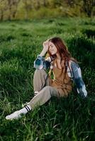 un' giovane, bellissimo donna salotti su il verde erba nel il parco indossare sneaker pantaloni e un' plaid camicia e sembra su in il ambientazione sole foto