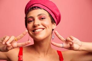 giovane atletico donna con un' corto taglio di capelli e viola capelli nel un' rosso superiore e un' rosa cappello con un atletico figura sorrisi e smorfie guardare a il telecamera su un' rosa sfondo foto