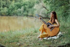 hippie donna giocando chitarra sorridente e cantando canzoni nel natura seduta su un' plaid di il lago nel il sera nel il raggi di il ambientazione sole. un' stile di vita nel armonia con il corpo e natura foto