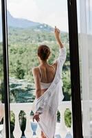 ritratto di un' bellissimo donna nel un' bianca camicia ammira il verde natura su il balcone soleggiato giorno foto
