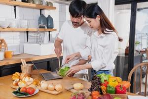 giovane asiatico coppia cucinando con frutta e verdure e utilizzando il computer portatile nel il cucina per cucinare cibo insieme entro il famiglia felicemente, famiglia concetto. foto