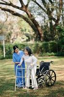 asiatico attento badante o infermiera e il contento paziente nel un' sedia a rotelle siamo a piedi nel il giardino per Aiuto e incoraggiare e riposo il tuo mente con verde natura. Aiuto supporto te stesso per imparare per camminare. foto