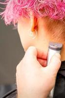 indietro Visualizza di parrucchiere mano rasatura nuca e collo con elettrico trimmer di giovane caucasico donna con corto rosa capelli nel bellezza salone. foto