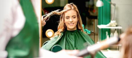 parrucchiere utilizzando arricciatura pinza riccioli lungo Marrone capelli su il giovane caucasico ragazza nel un' bellezza salone. foto