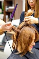 Due parrucchieri utilizzando arricciatura ferro su i clienti lungo Marrone capelli nel un' bellezza salone. foto
