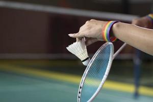 badminton giocatore indossa arcobaleno braccialetti e Tenere racchetta e bianca volano nel davanti di il netto prima servendo esso per giocatore nel un altro lato di il Tribunale, concetto per lgbt persone attività. foto