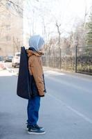 un' adolescenziale ragazzo nel un' giacca e cappello passeggiate solo con un' chitarra nel un' Astuccio. bambino musicista perso nel il città foto