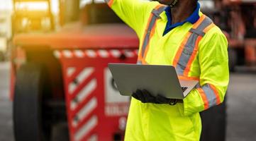 vicino su tecnico bacino lavoratore nel protettivo sicurezza tuta uniforme e con elmetto protettivo e uso il computer portatile computer a carico contenitore spedizione magazzino. mezzi di trasporto importare, esportare logistica industriale foto