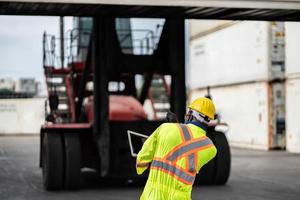 africano tecnico bacino lavoratore nel protettivo sicurezza tuta uniforme e con elmetto protettivo e uso digitale tavoletta a carico contenitore spedizione magazzino. mezzi di trasporto importare, esportare logistica industriale foto