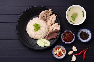 hainanese pollo riso su un' nero di legno tavolo sfondo al vapore pollo riso con pollo la minestra, salse condimenti asiatico cibo Tailandia Cina Singapore, superiore Visualizza foto