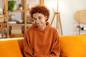 bellissimo africano americano ragazza con afro acconciatura sorridente seduta su divano a casa interno. giovane africano donna con Riccio capelli ridendo. la libertà felicità spensierato contento persone concetto. foto