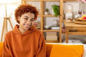 bellissimo africano americano ragazza con afro acconciatura sorridente seduta su divano a casa interno. giovane africano donna con Riccio capelli ridendo. la libertà felicità spensierato contento persone concetto. foto