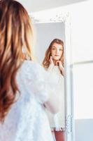 bellezza trucco mattina rutina amore te stesso concetto. giovane adolescenziale ragazza guardare a riflessione nel specchio. giovane positivo donna indossare bianca vestito in posa nel luminosa leggero camera contro bianca parete. foto