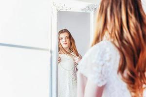 bellezza trucco mattina rutina amore te stesso concetto. giovane adolescenziale ragazza guardare a riflessione nel specchio. giovane positivo donna indossare bianca vestito in posa nel luminosa leggero camera contro bianca parete. foto