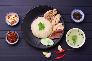 hainanese pollo riso su un' nero di legno tavolo sfondo al vapore pollo riso con pollo la minestra, salse condimenti asiatico cibo Tailandia Cina Singapore, superiore Visualizza foto