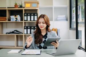 bellissimo donna digitando su tavoletta e il computer portatile mentre seduta a il Lavorando di legno tavolo moderno ufficio foto