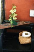 fazzoletto di carta nel un' cestino e orchidea fiori su di legno mensola sospeso su arancia parete decorazione nel bagno Vintage ▾ stile foto