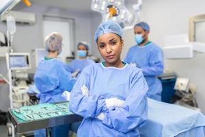 avvicinamento di un' africano americano chirurgo donna guardare a telecamera con colleghi l'esecuzione nel sfondo nel operazione camera. il concetto di medicina foto