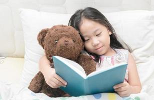 carino poco asiatico ragazza nel camicia da notte lettura un' libro con orsacchiotto orso su letto. foto