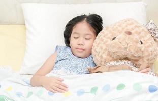 adorabile poco ragazza addormentato su il letto con sua orsacchiotto orso foto