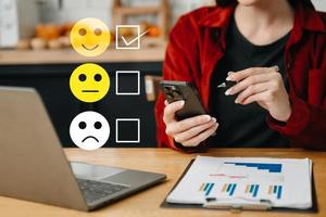 cliente servizio valutazione concetto. donna mostrare viso Sorridi emoticon mostrare su virtuale schermo a partire dal mano.guardando a inteligente Telefono, tavoletta e il computer portatile nel ufficio foto