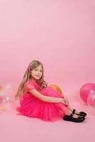 sorridente sette anni ragazza vestito nel un' alla moda vestito si siede su un' rosa sfondo circondato di palloncini. celebrare il tuo compleanno foto