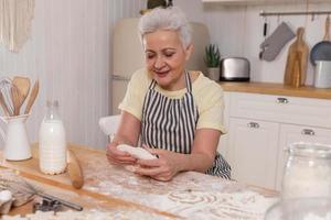 contento anziano donna cucinando nel cucina. elegante più vecchio maturo grigio dai capelli signora nonna impastare Impasto infornare biscotti. vecchio nonna cucinare fatti in casa cibo. domestico casalinga lavori di casa concetto. foto