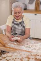 contento anziano donna cucinando nel cucina. elegante più vecchio maturo grigio dai capelli signora nonna impastare Impasto infornare biscotti. vecchio nonna cucinare fatti in casa cibo. domestico casalinga lavori di casa concetto. foto